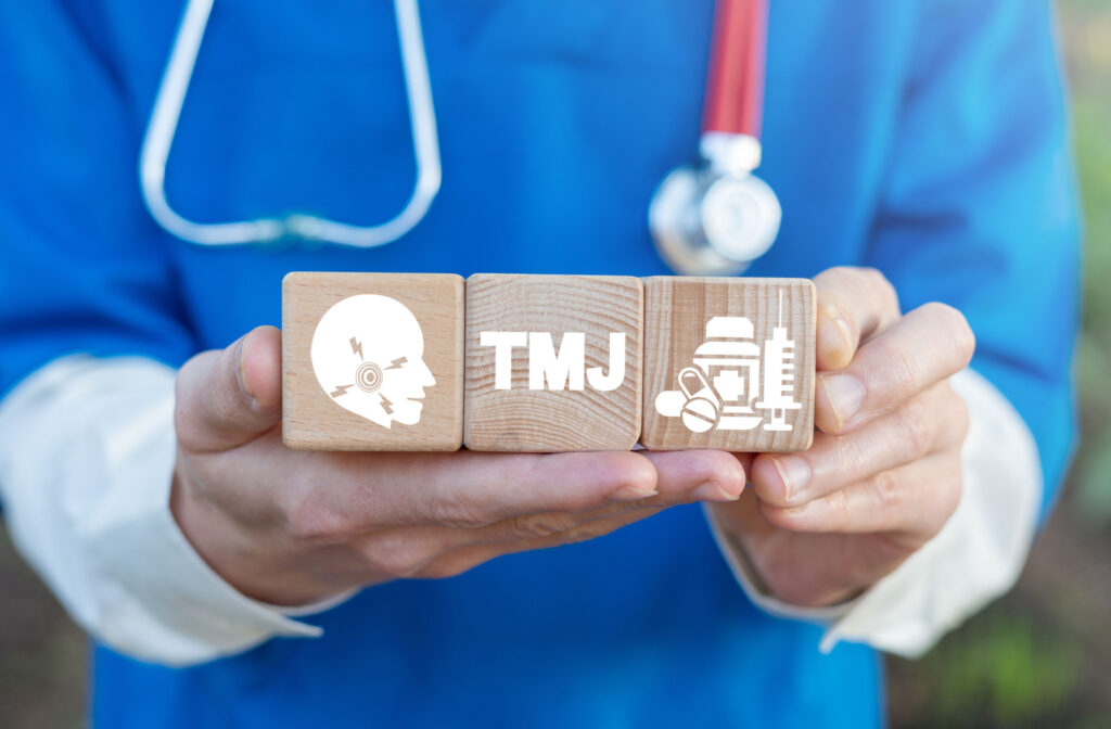 a dentist wearing scrubs holds up three wooden blocks with images on them. one shows an illustrated cross section of a jaw, one shows a pain medication and the other says "tmj"