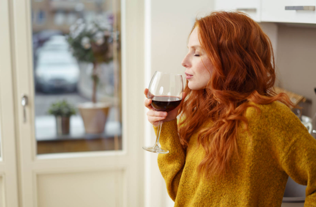 A young red-haired woman taking a whiff of a glass of red wine before drinking it.