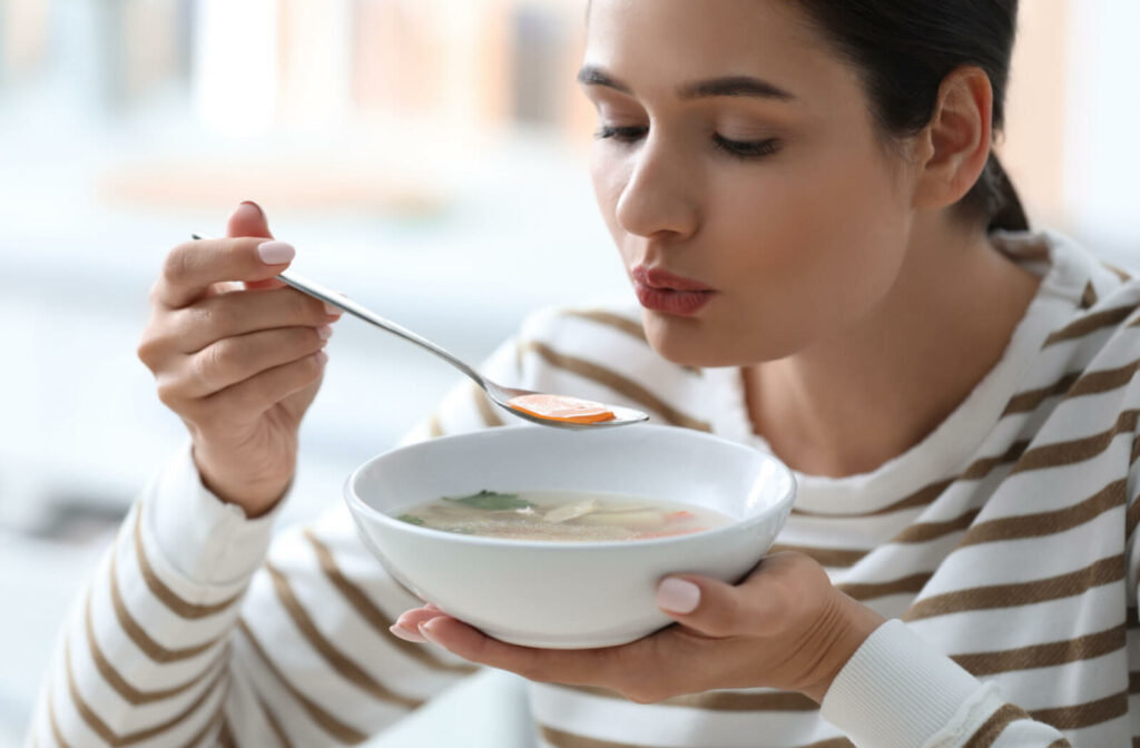 A woman in a striped shirt carefully savors each spoonful of vegetable soup during her post-wisdom tooth extraction recovery