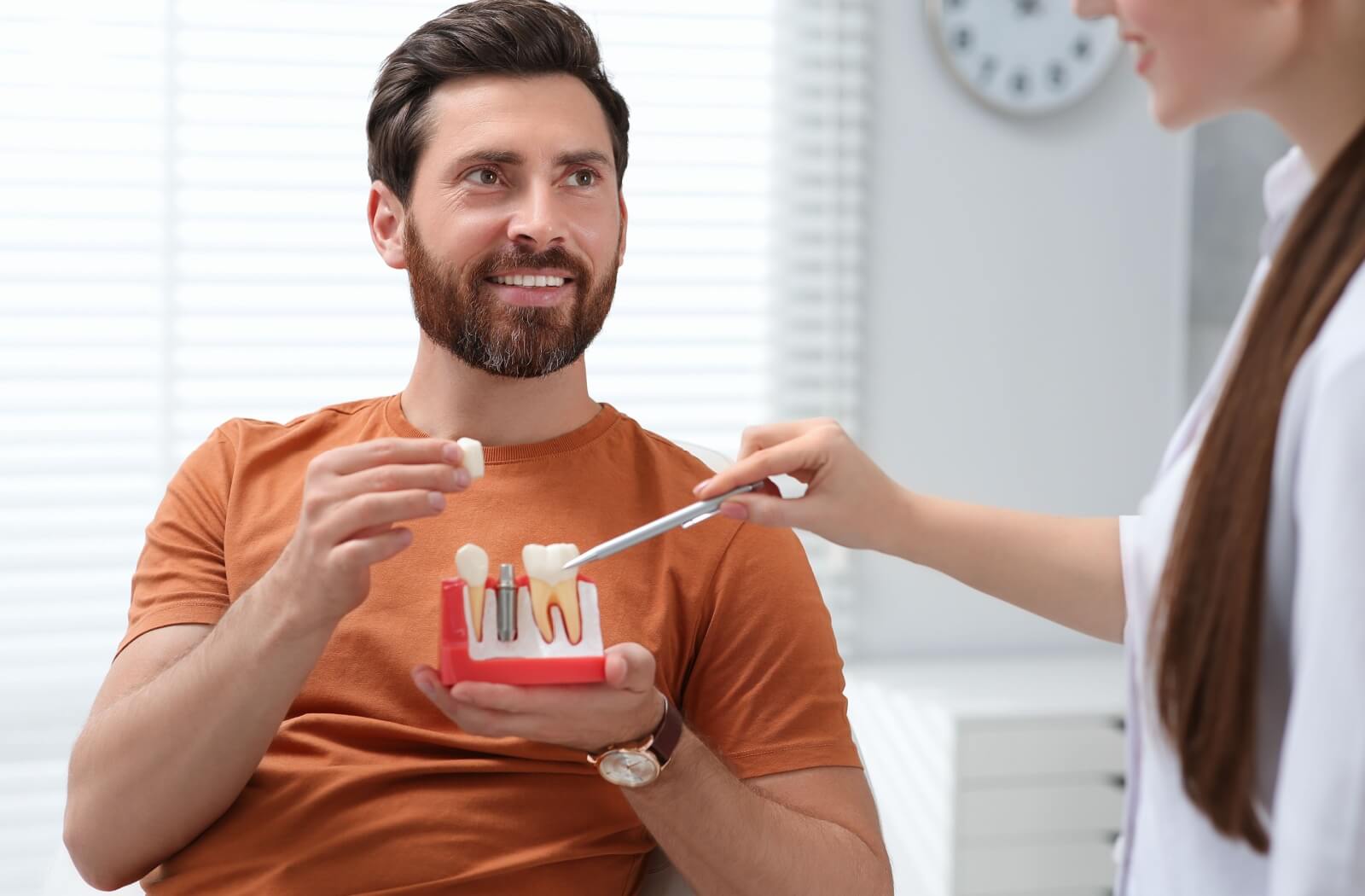 A dentist points to a partial model of teeth and gums with a post that has screws coming out of the top, explaining how a dental implant works. A patient holds the model in their left hand while holding the top of the artificial tooth in their right.