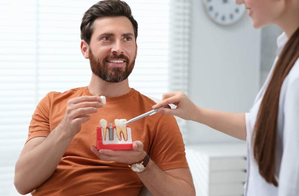 A dentist points to a partial model of teeth and gums with a post that has screws coming out of the top, explaining how a dental implant works. A patient holds the model in their left hand while holding the top of the artificial tooth in their right.