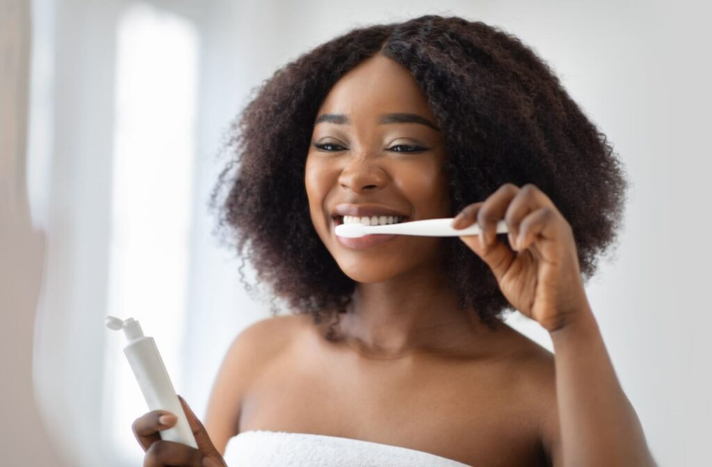 A young woman brushing her teeth in front of the mirror in a sunlit bathroom.