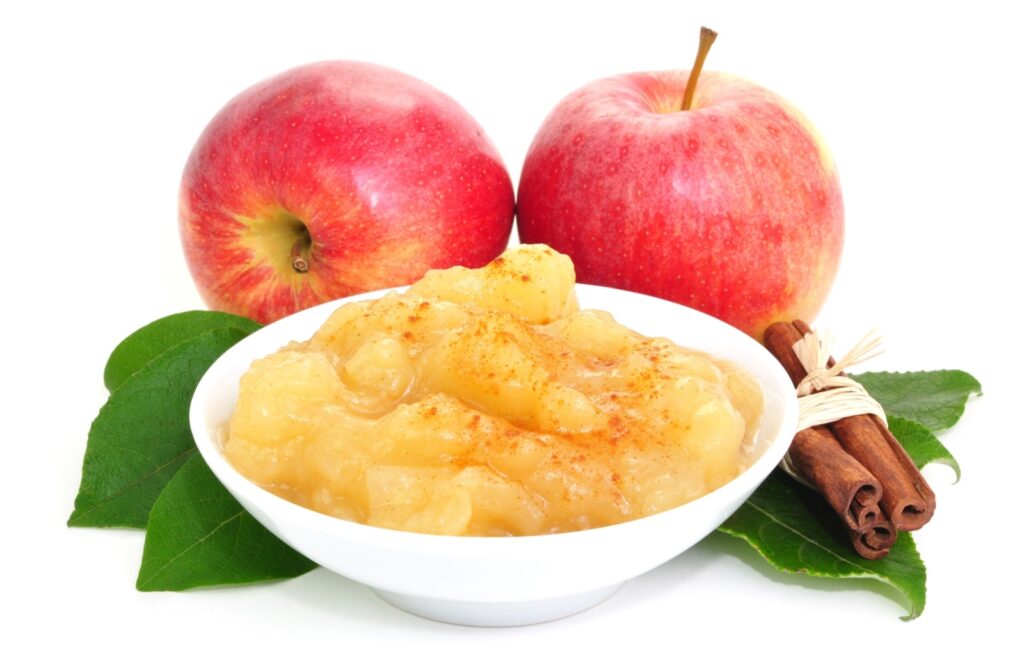 Close-up of apple sauce in a white dish sprinkled with cinnamon. Two red apples, green leaves, and three sticks of cinnamon surround the dish.