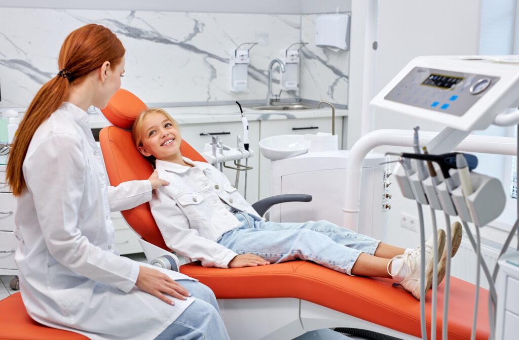 A smiling child sitting in an orange dental exam chair in a brightly lit room as a dentist explains the sealant process to them.
