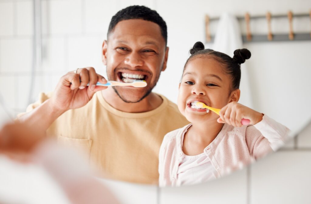 A parent and their child brushing their teeth together in front of a mirror in a brightly lit bathroom.