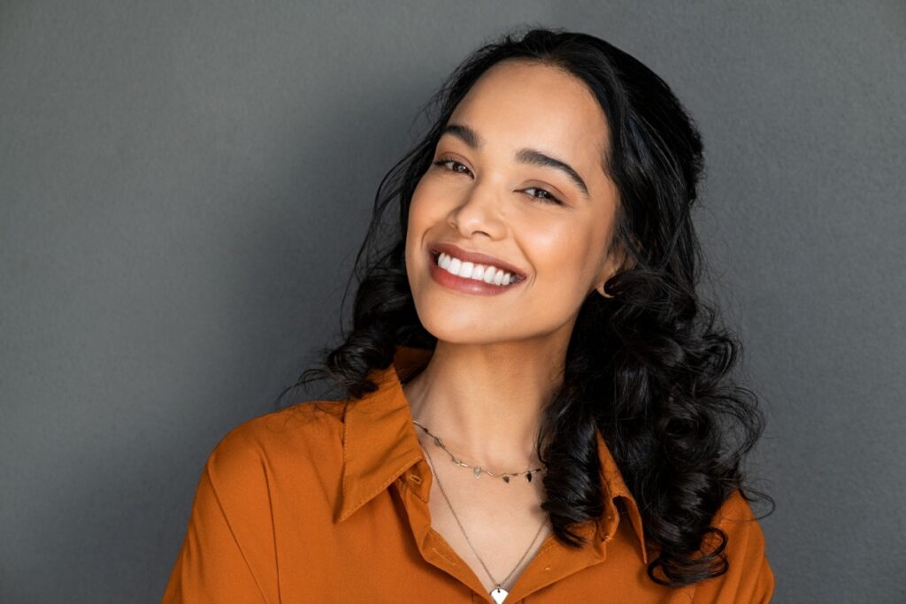 Smiling woman with curly hair wearing an orange shirt, posing against a gray background.