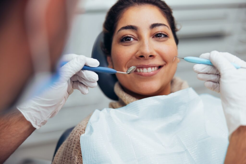 Woman sitting in a dental chair smiling while a dentist holds dental instruments near her mouth.