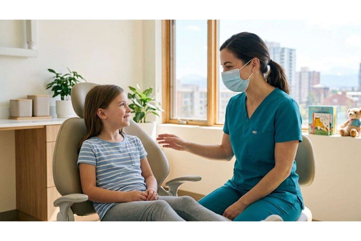 A friendly female pediatric dentist in blue scrubs and a face mask sitting and talking with a young girl in a dental chair in a bright, welcoming office at Sunshine Dental.